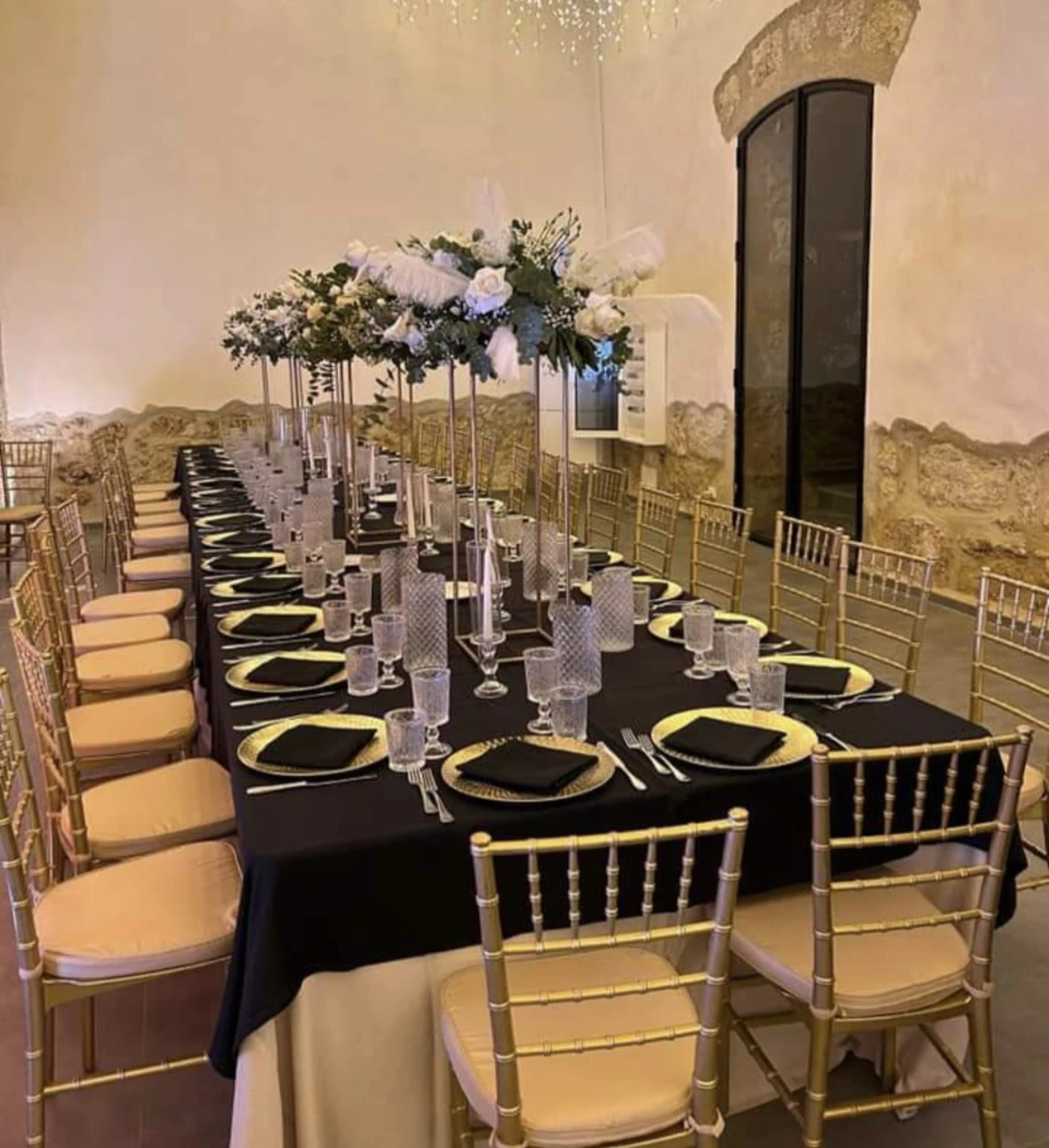 Tavolata nera con sedie dorate e alzate di fiori bianchi, nella sala in pietra